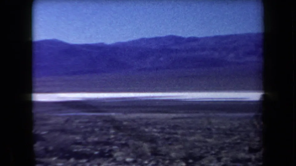 Wide 8mm view of an Arizona desert landscape in 1958, with rocky ground, a pale flat strip, distant mountains, and no clear people visible.