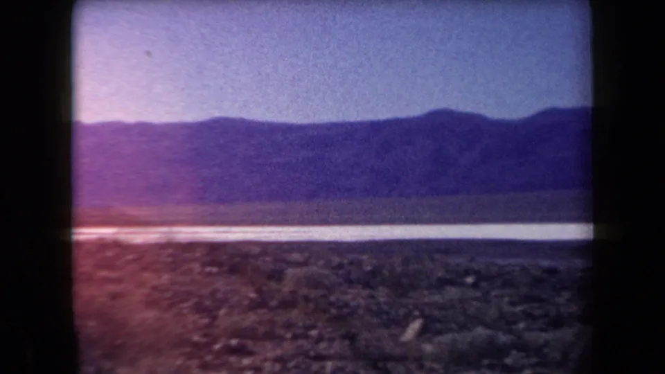 Faded 8mm frame of the Arizona desert, with rocky foreground, a bright pale flat, and mountains under a lightly tinted sky.