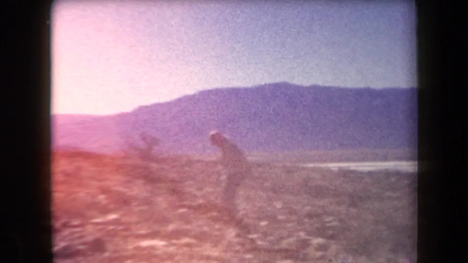 Soft, faded 1958 Arizona 8mm frame with two people moving low over rocky desert ground, backed by a mountain ridge.