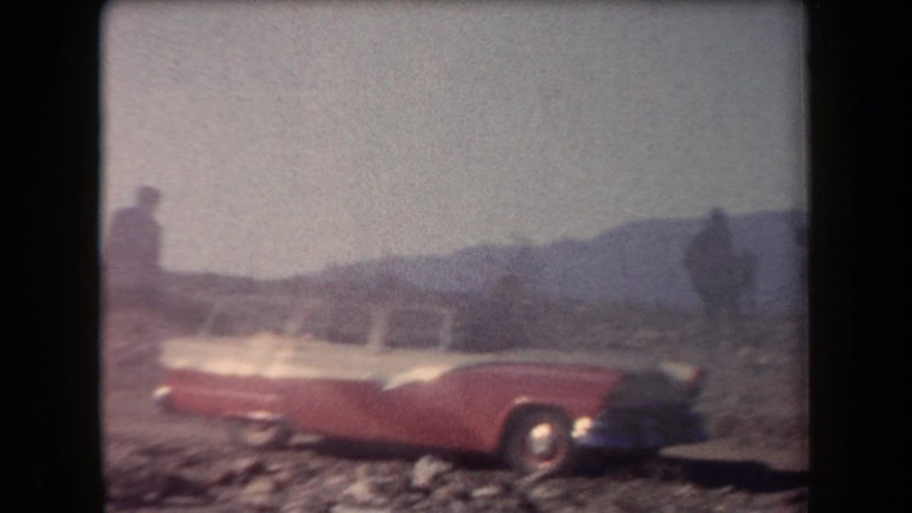 Red-and-white station wagon parked on rocky desert ground with mountains behind it.