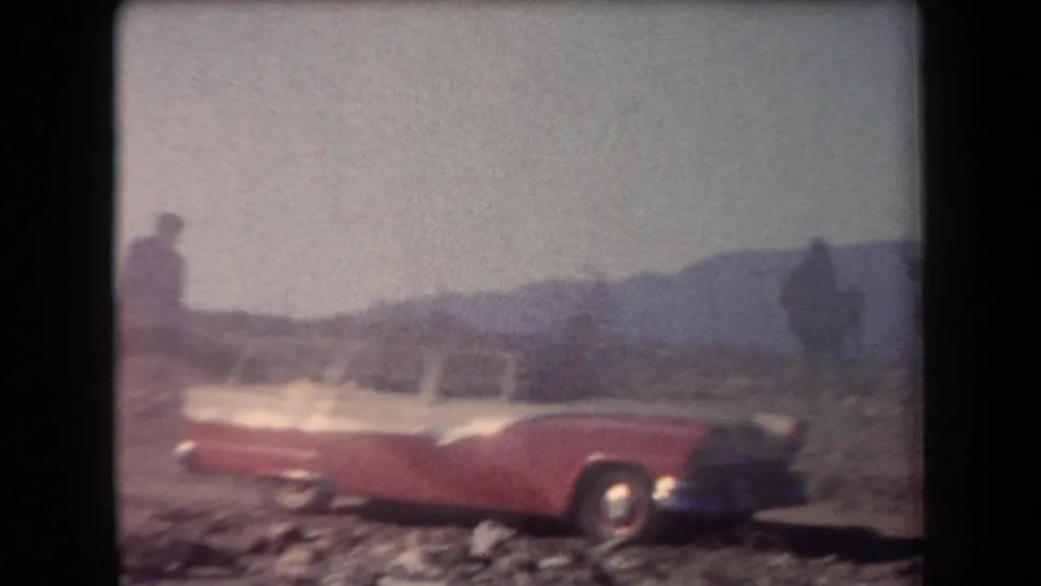A red-and-white mid-century station wagon sits in the Arizona desert, with distant figures and mountains visible through faded 8mm color.