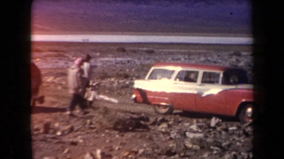 Several travelers walk near a red-and-white station wagon on rocky Arizona desert terrain, with mountains and a pale horizontal strip beyond.