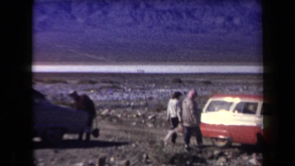 In a 1958 Arizona desert stop, travelers walk near a red-and-white station wagon and a darker car against a mountain backdrop.