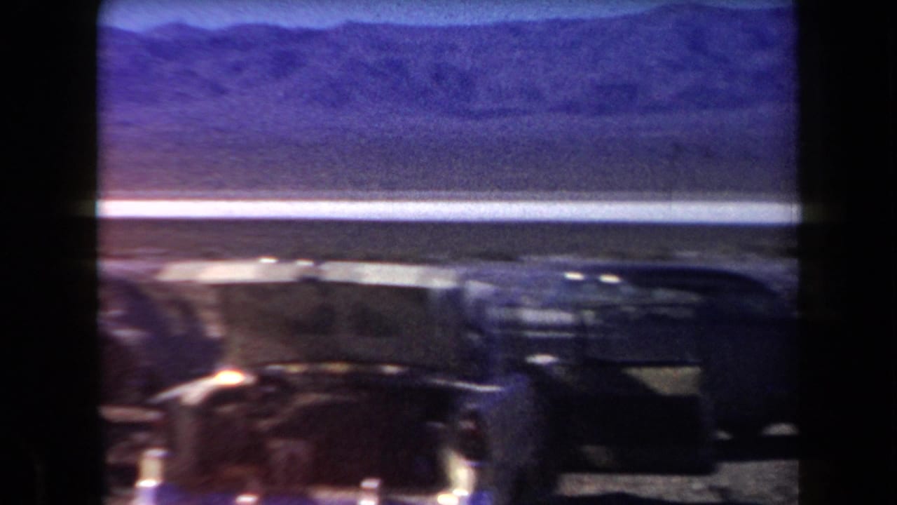 A dark car passes or fills the foreground with desert flats and mountains behind it.