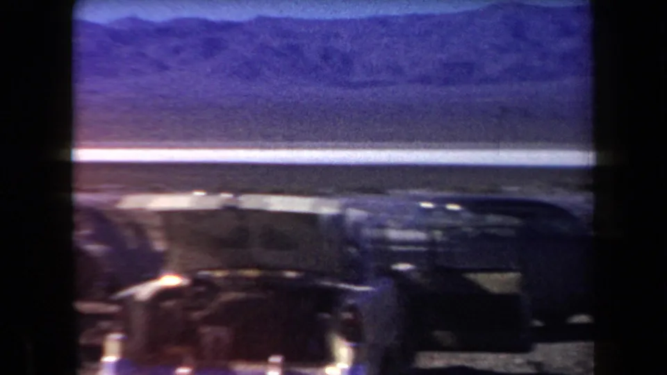 A classic car moves through or near the foreground while the camera faces a pale desert flat and mountain range in Arizona.