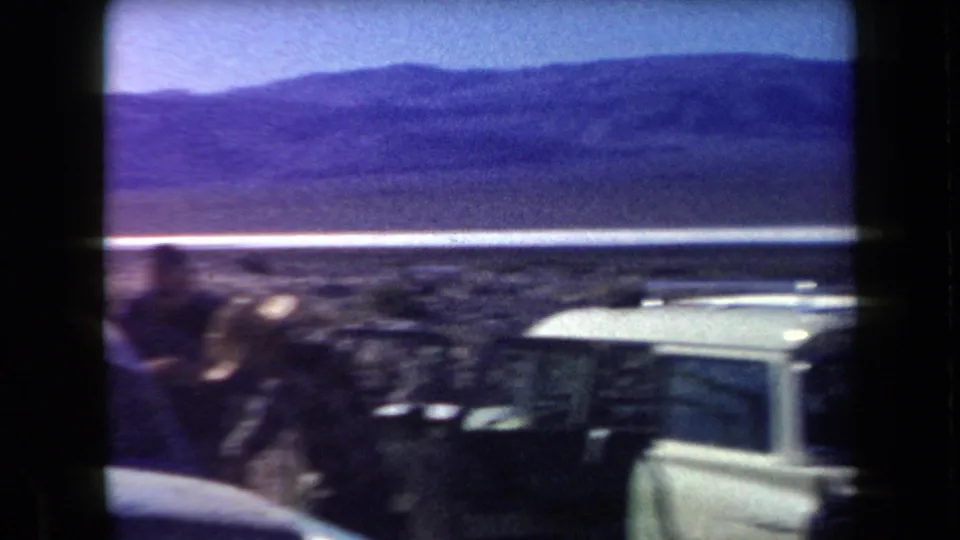 A blurred traveler and light-colored car are seen during a handheld desert roadside shot with mountains beyond.