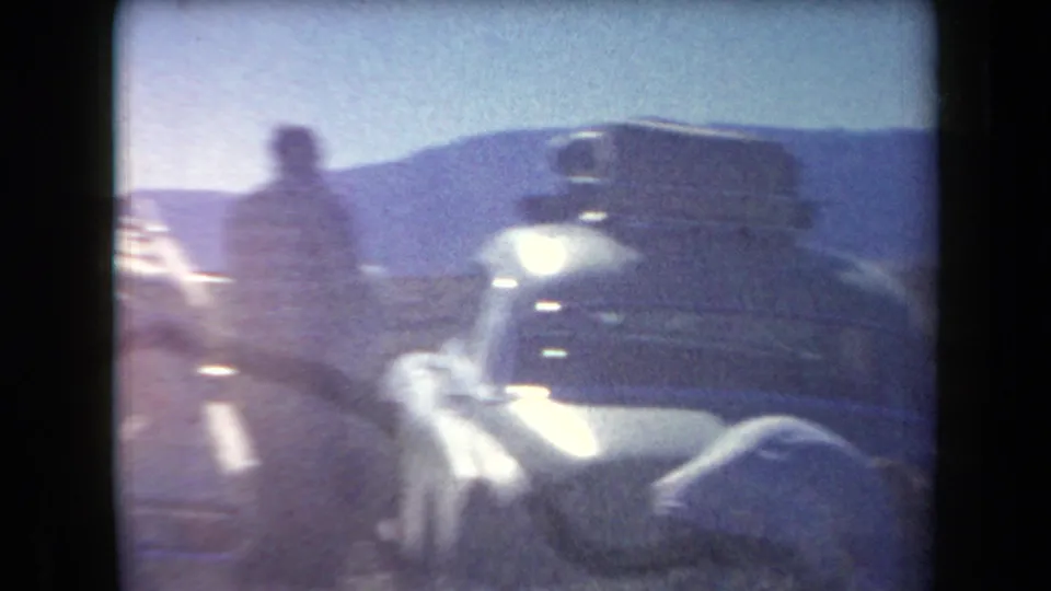 A light-colored classic car, possibly carrying roof luggage, sits near a blurred adult figure in the 1958 Arizona desert road-trip footage.