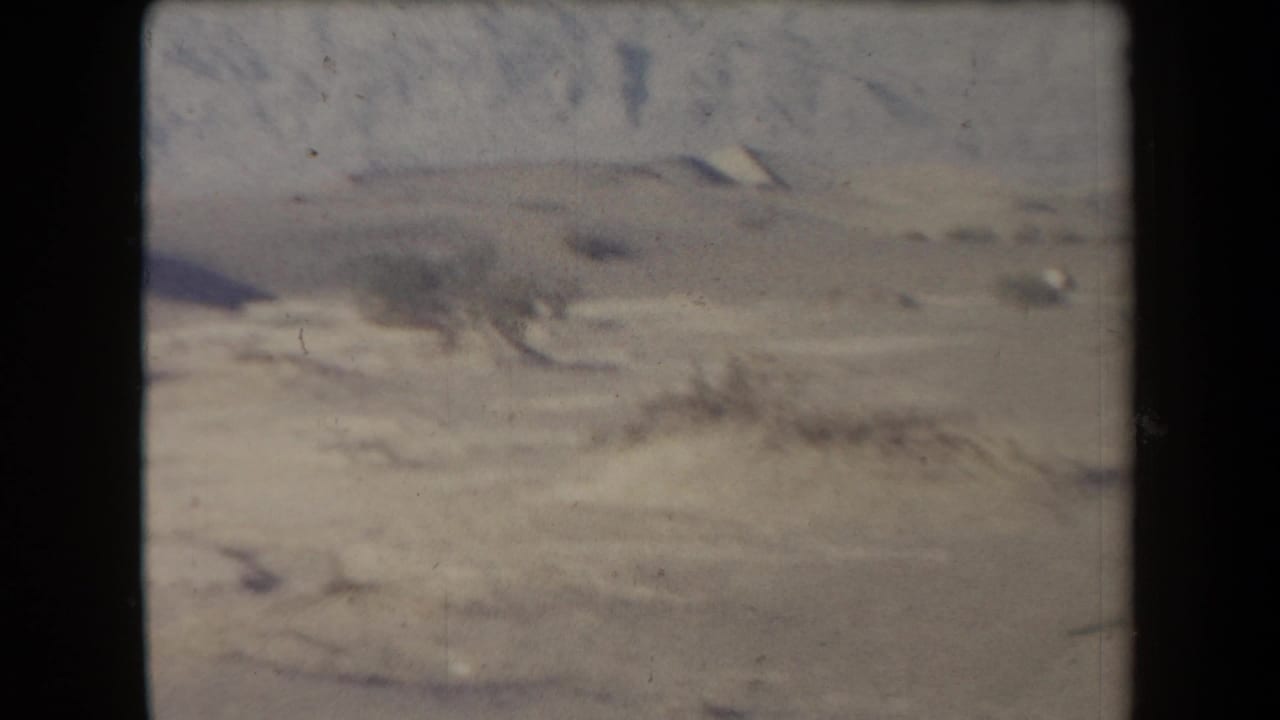 Pale desert ground with distant hills and sparse scrub under a hazy sky.