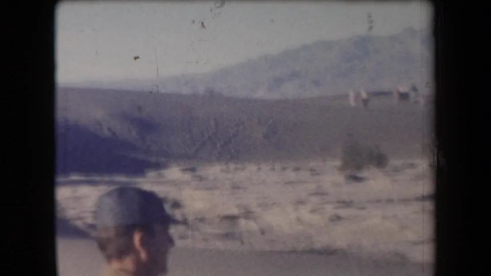 Arizona 8mm travel frame showing a man in a cap partly in foreground, with desert scrub, hills, and distant tourists behind him.