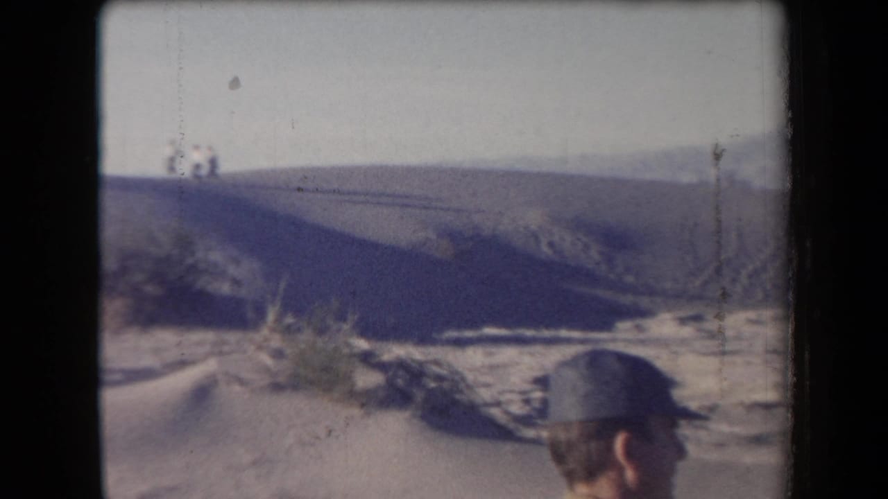 1958 Arizona home-movie frame showing a capped observer near desert dunes, scrub, and distant mountains.