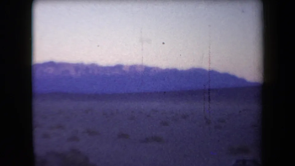 1958 Arizona home-movie frame with distant figures on a desert flat beneath a blue-purple mountain range.