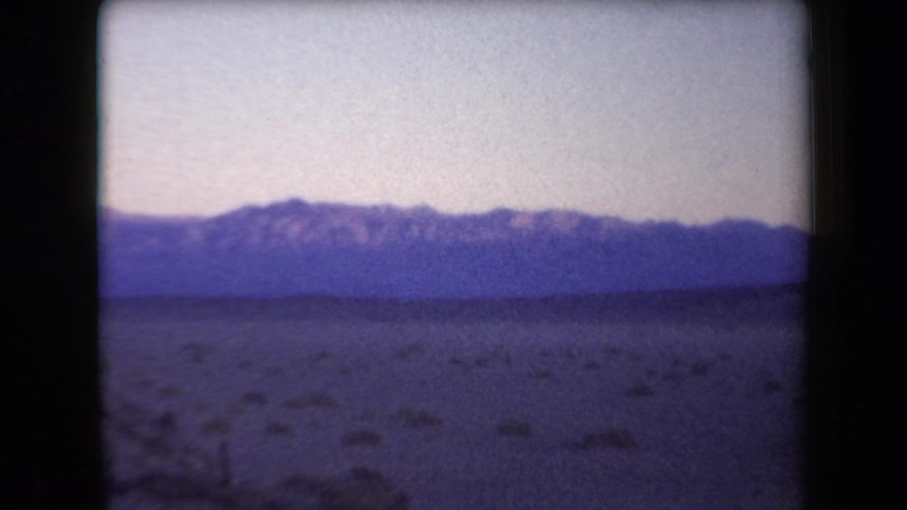 Arizona 1958 8mm frame showing a sandy desert foreground and distant purple-blue mountains beneath a pale evening sky.