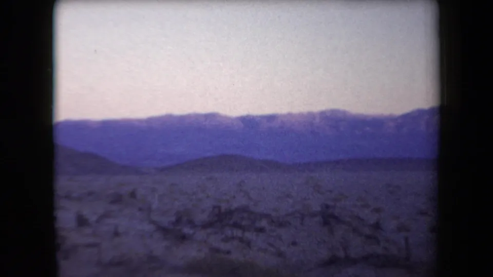 Arizona 1958 home-movie frame with low desert hills, sparse brush, and a soft pale sky near dusk.