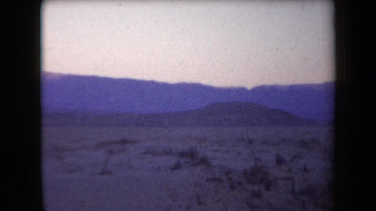 1958 Arizona 8mm frame showing a dry desert foreground, low scrub, and blue-purple mountain layers beyond.