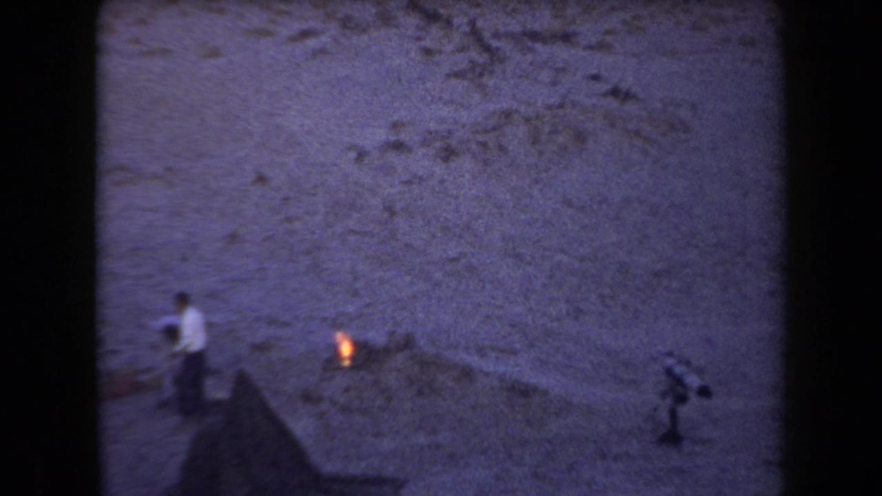 1958 Arizona home-movie frame showing a person near a small campfire, tent edge, and rocky desert cliff at dusk.