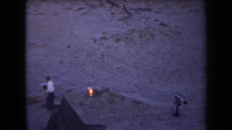 1958 Arizona home-movie frame showing a person near a small campfire, tent edge, and rocky desert cliff at dusk.