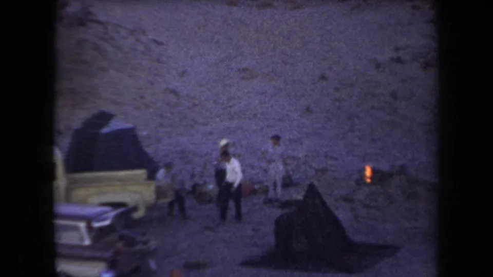 1958 Arizona camping scene with people near a light-colored pickup, a small fire, and a rocky desert backdrop.