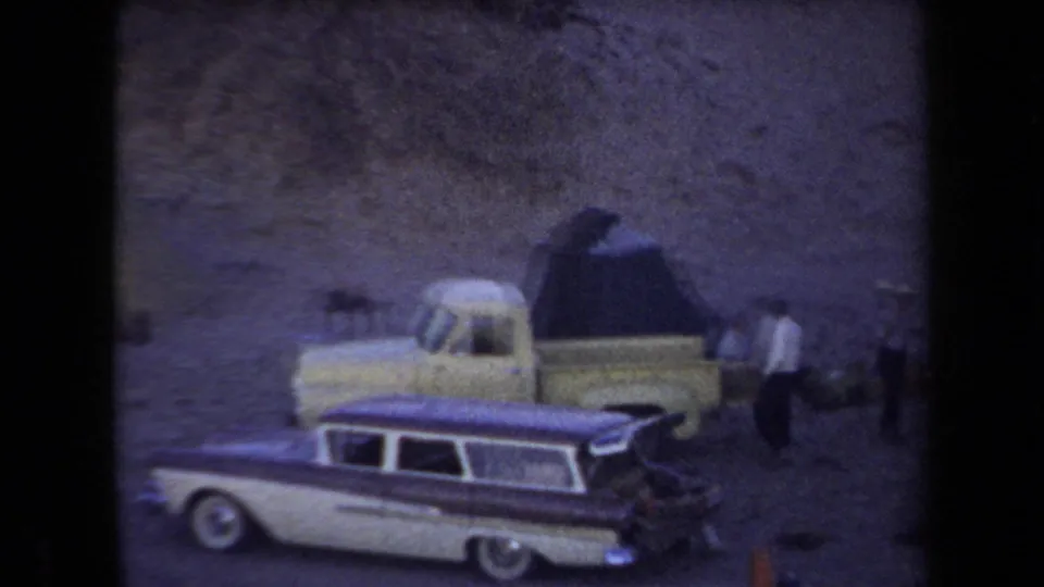 1958 Arizona home-movie frame with a station wagon, pickup truck, tent, and people moving around a desert camp.