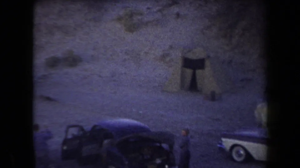 1958 Arizona desert campsite frame showing a dark car with an open door, a partial station wagon, and camp gear near a rock wall.