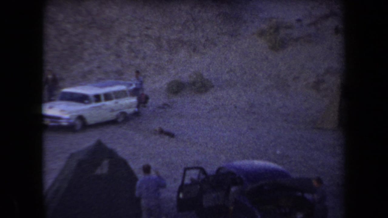 1958 Arizona 8mm frame showing a white station wagon, tent camp, and people on sandy desert terrain.