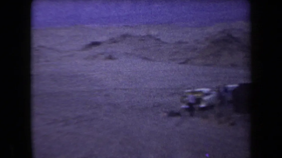 Grainy 1958 Arizona desert frame with low sandy ridges and partial campsite objects near the foreground.