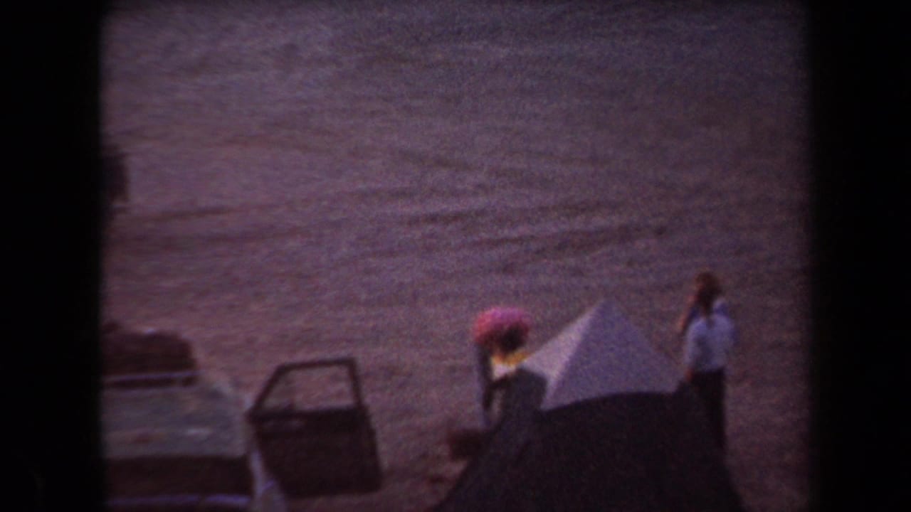 1958 Arizona home-movie frame with two people standing near classic cars on dry open ground.