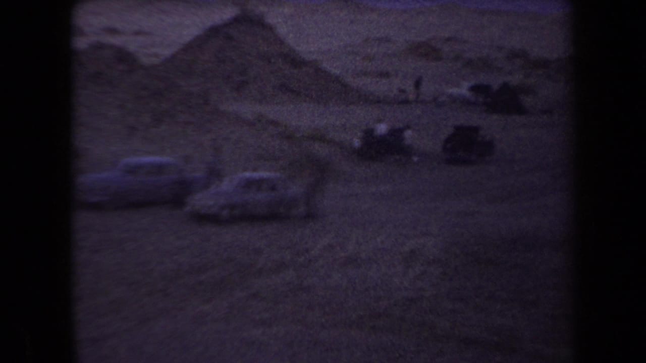 Arizona 1958 home-movie frame showing parked cars and small visitors near sandy desert mounds at dusk.