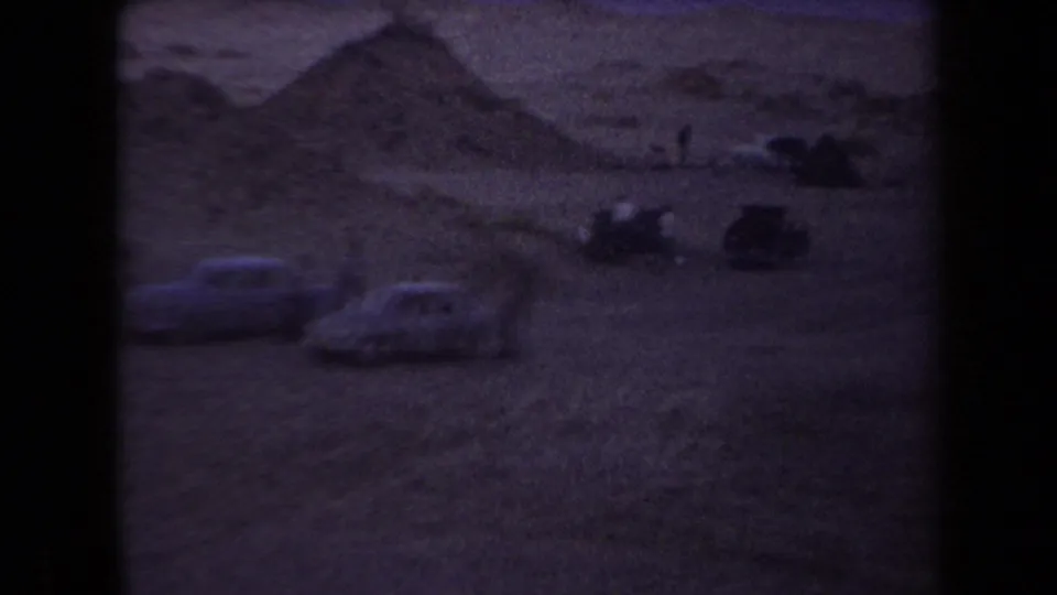Arizona 1958 home-movie frame showing parked cars and small visitors near sandy desert mounds at dusk.