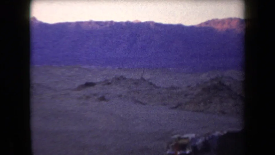 Grainy Arizona desert frame with low rocky ground, mountain horizon, and dark foreground edge from the handheld pan.