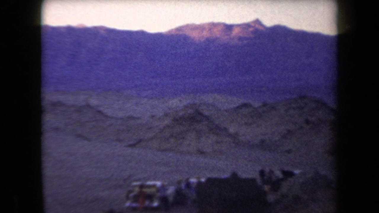 1958 Arizona 8mm frame showing a remote desert campsite beneath mountains at dusk, with tents, vehicles, and people near camp.
