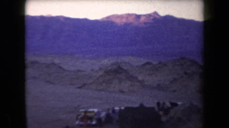 1958 Arizona 8mm frame showing a remote desert campsite beneath mountains at dusk, with tents, vehicles, and people near camp.