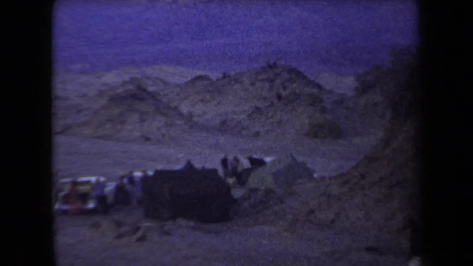 1958 Arizona home-movie frame of a tent and parked vehicles among rocky desert hills, with people silhouetted near the campsite.