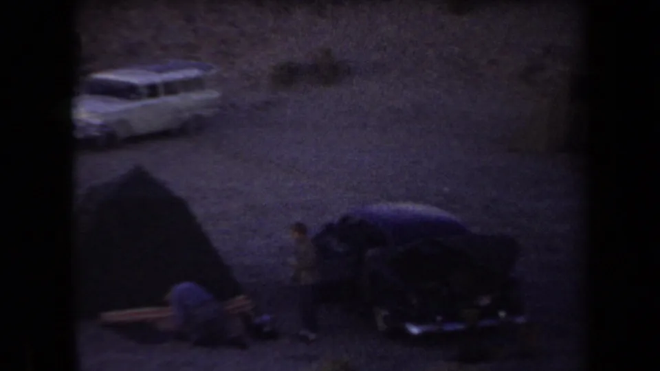 1958 Arizona home-movie frame showing cars, a tent, and people at a desert campsite surrounded by rocky hills at dusk.