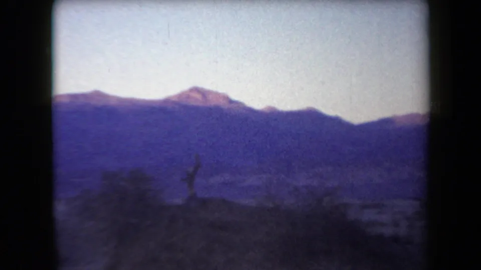 1958 Arizona 8mm frame showing a blue-toned mountain range beyond shadowed desert brush and open sandy ground.