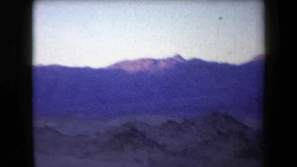 1958 Arizona landscape frame with open sandy desert, low shrubs, distant purple mountains, and aged 8mm color shift.