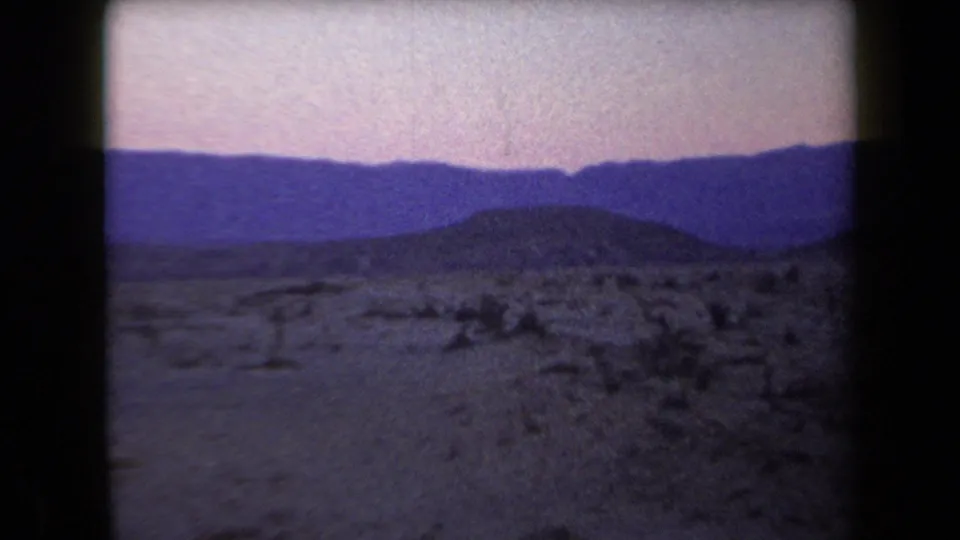 1958 Arizona home-movie frame showing low desert hills, scattered shrubs, distant mountains, and a pink-purple twilight sky.