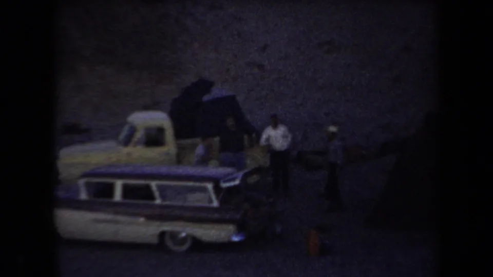 Arizona 1958 8mm frame showing a station wagon at a desert campsite with gear or a tent and distant standing figures.