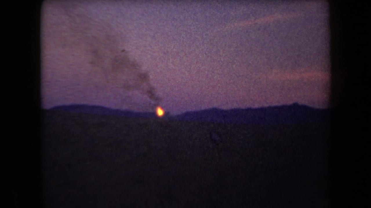 Distant orange fire glows behind a dark mountain ridge as smoke rises into a purple night sky.