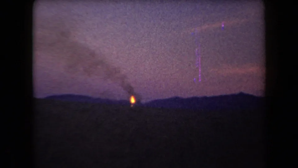 1958 Arizona home-movie frame with a distant orange flame, smoke plume, silhouetted hills, and visible low-light film grain.