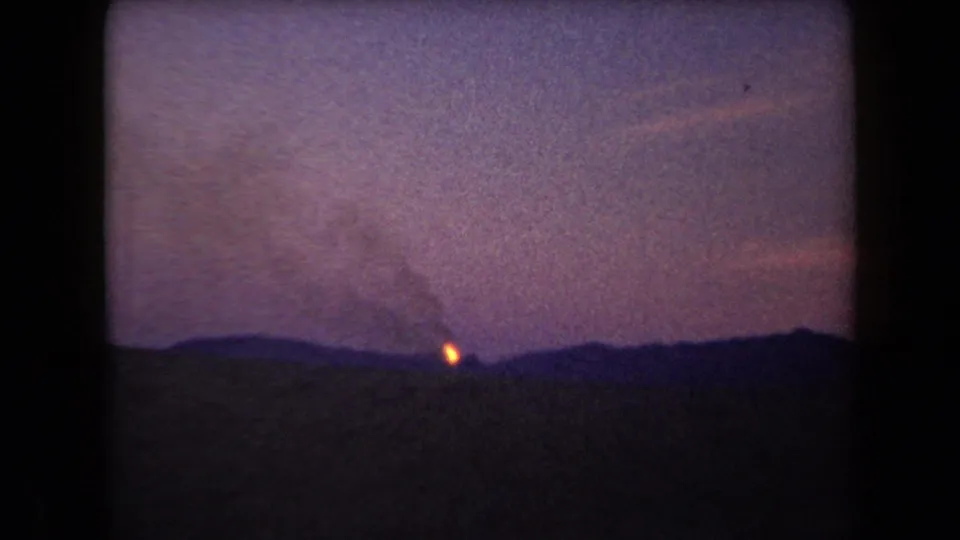 1958 Arizona 8mm frame with a distant wildfire glow behind a dark ridge, smoke overhead, and soft grain from low-light filming.