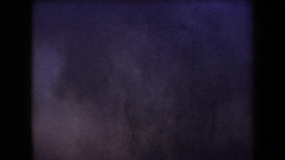 Grainy 8mm low-light frame with purple dusk tint, soft exposure, and indistinct ground or smoke forms.
