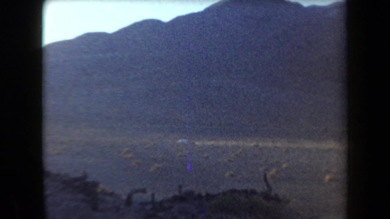 1958 Arizona home-movie frame showing a barren desert basin below dark mountain slopes, with no people visible.