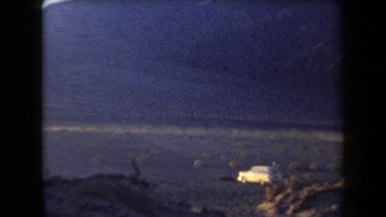 A pale car appears parked in the Arizona desert in 1958, set against brush, rocky ground, and distant mountains.