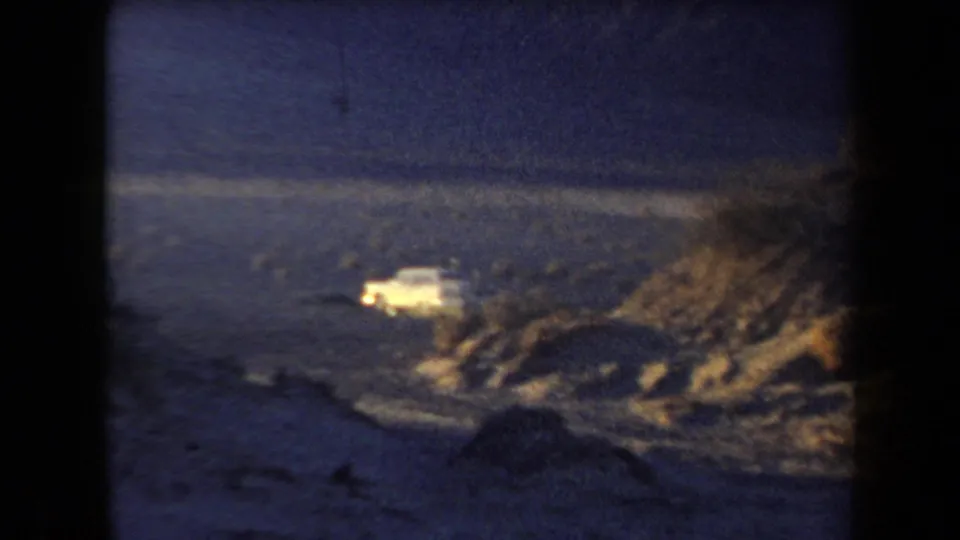 1958 Arizona 8mm frame with a pale car beside a rocky desert slope and distant mountain backdrop.