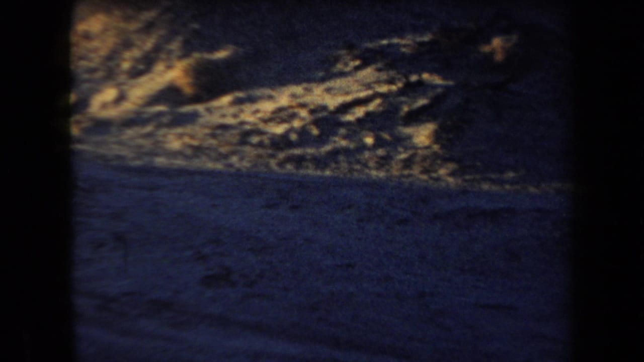 Low-light 1958 Arizona 8mm frame showing dark sand and uneven warm highlights, with no clear people visible in this still.