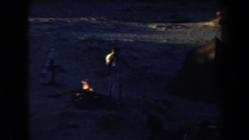 1958 Arizona 8mm home-movie frame showing a sandy nighttime campsite with a campfire, distant silhouetted people, and a dark tent.