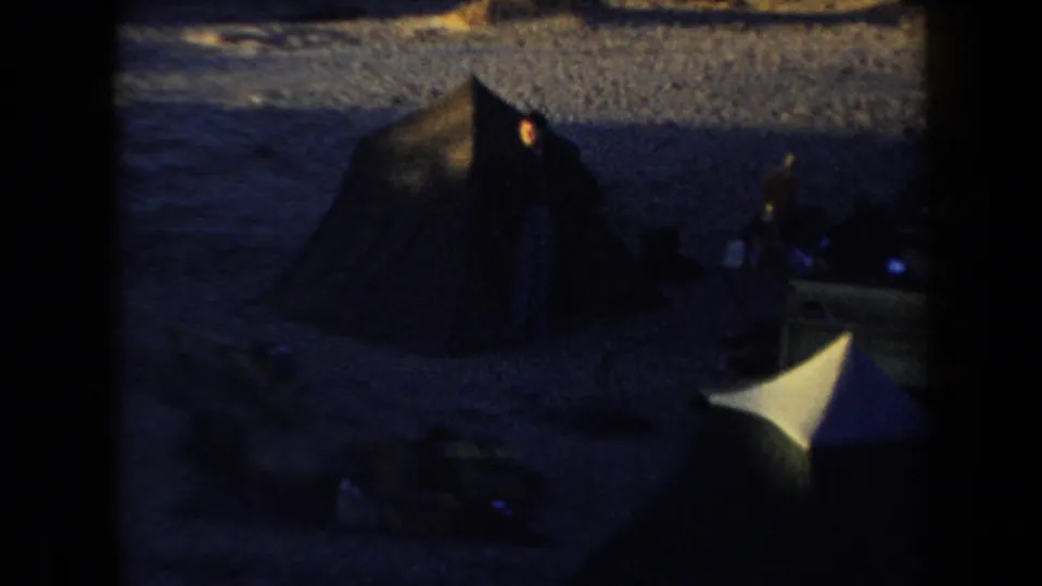 1958 Arizona campsite frame with dark tents, scattered camping gear, sandy ground, and a small warm fire glow in low light.