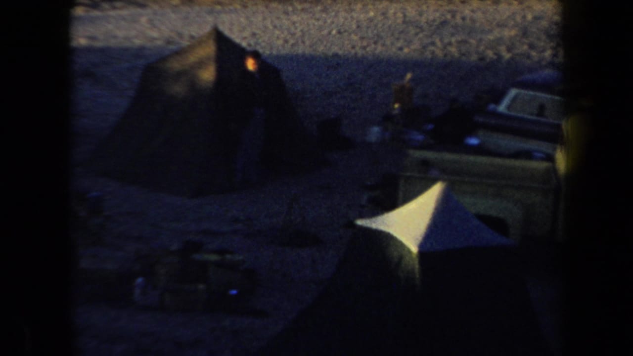 Dark campsite with canvas tents, parked vehicles, gravel ground, and a small indistinct person.