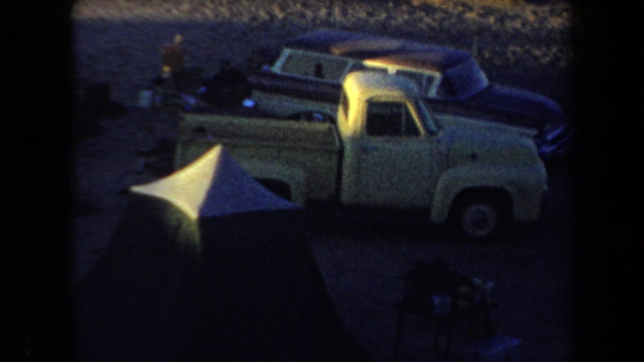 A light pickup truck and dark classic cars sit near a tent in low-light 1958 Arizona camping footage.