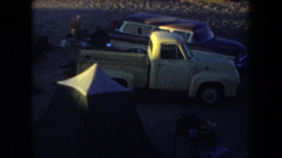 A light pickup truck and dark classic cars sit near a tent in low-light 1958 Arizona camping footage.